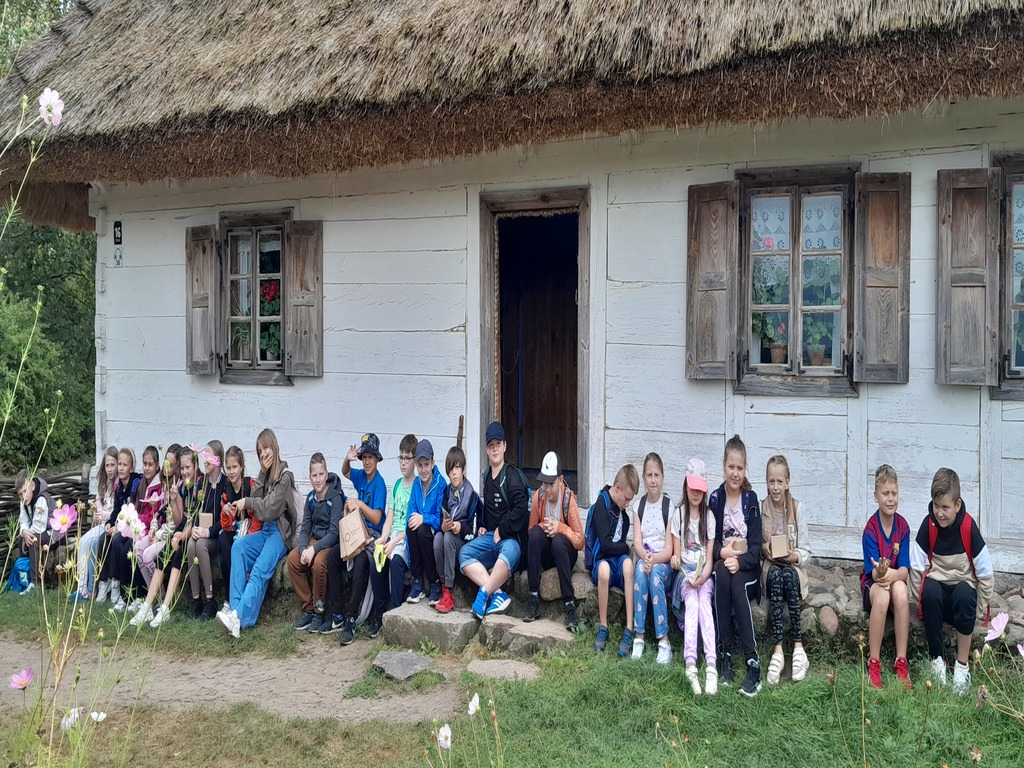 Uczestnicy wycieczki siedzą przed chatą w skansenie